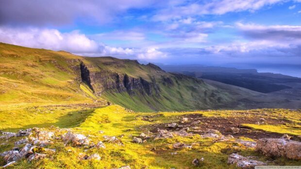 Rolling hills and rugged cliffs of Isle of Skye covered in green grass under a cloudy blue sky