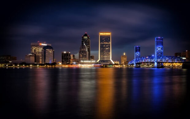 Nighttime cityscape of Jacksonville with illuminated skyscrapers and a lit bridge reflecting on the river