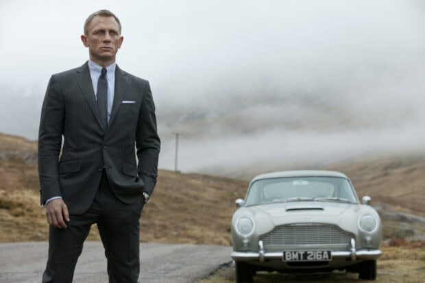 James Bond standing in a suit near a classic car on a misty rural road