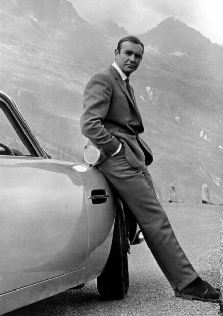 Sean Connery leaning against a classic car in a mountainous landscape with a suit