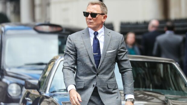 James Bond in a gray suit and sunglasses walking confidently near a vintage car in the city