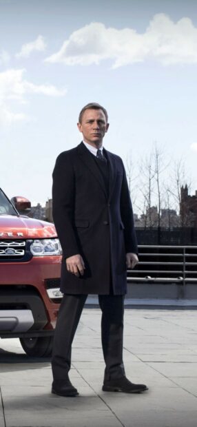 James Bond standing next to a red Land Rover wearing a black coat outdoors