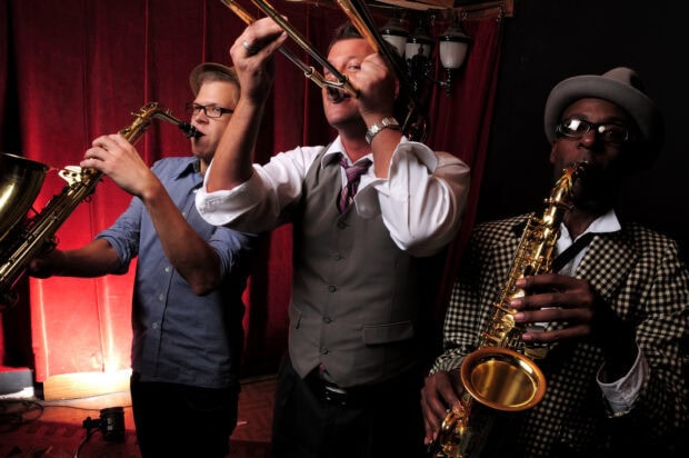 Three jazz musicians playing saxophone and trombone on stage with a red curtain background
