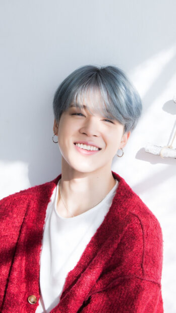 Smiling Jimin wearing red sweater and silver earrings in bright natural light