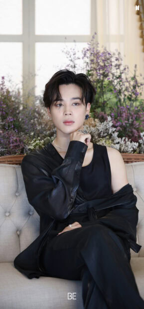 Jimin wearing black clothes sitting thoughtfully on a sofa surrounded by flowers