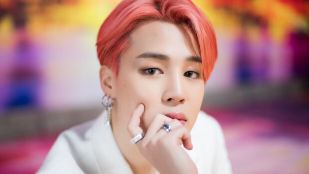 Jimin with pink hair and rings posing thoughtfully in a colorful setting