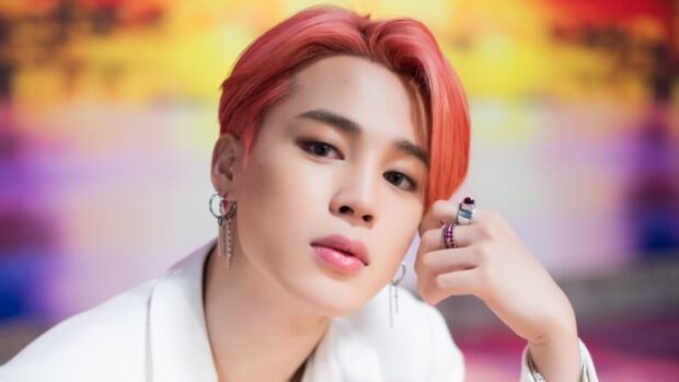Jimin with pink hair wearing multiple earrings and rings in a colorful blurred background