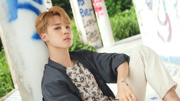 Jimin wearing a floral shirt and dark jacket sitting against a pillar outdoors in a casual pose