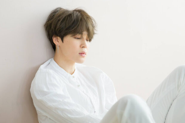 Jimin in a white outfit sitting calmly against a wall in a bright room