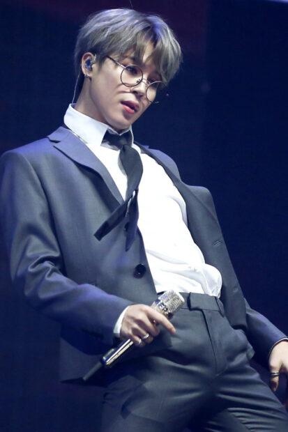 Jimin in a stylish suit performing on stage with glasses and holding a microphone