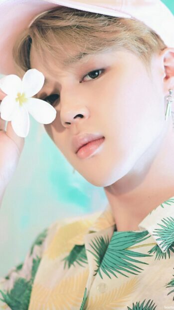 Jimin wearing a tropical shirt holding a white flower near his face in a close up portrait