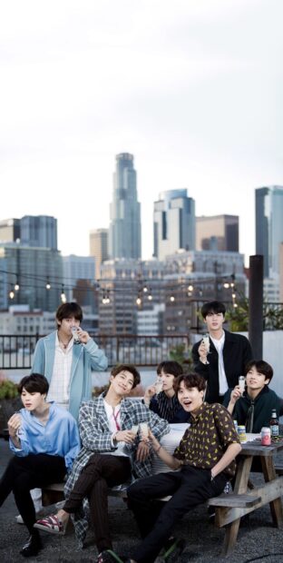 Jimin and BTS members enjoying drinks together on a rooftop with city skyline in the background