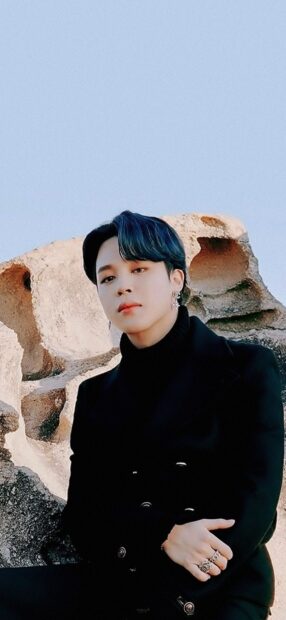 Jimin wearing a black coat and rings sitting against rock formations on a clear day