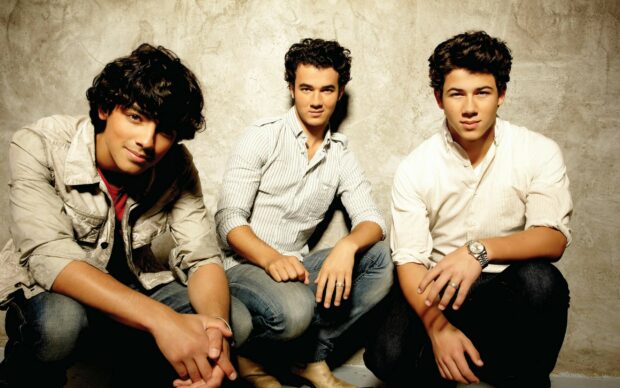 The Jonas Brothers sitting against a textured wall wearing casual clothes and looking at the camera