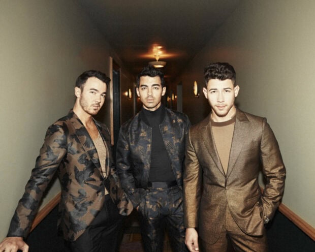 The Jonas Brothers standing together in patterned suits in a dimly lit hallway