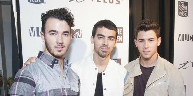 The Jonas Brothers standing together posing at an event with a branded backdrop behind them