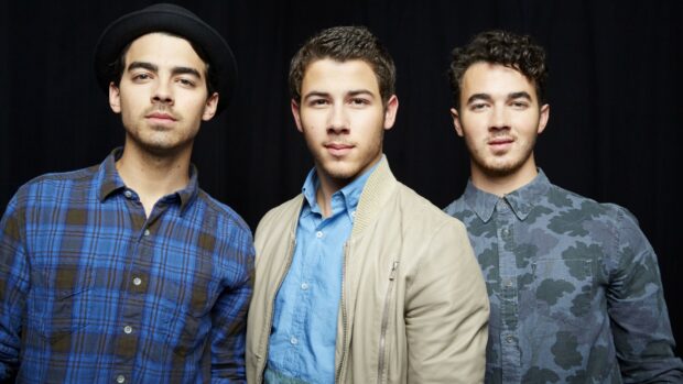 The Jonas Brothers band pose together against a dark background with casual outfits