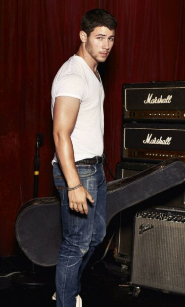 Nick Jonas wearing a white t shirt and jeans standing with a guitar case and amplifiers in the background