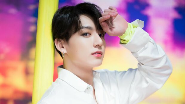 Jungkook wearing a white shirt posing with his hand near his face in a colorful setting