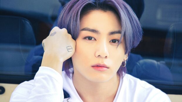 Jungkook with purple hair resting his hand on his face showing tattoos in a close up portrait