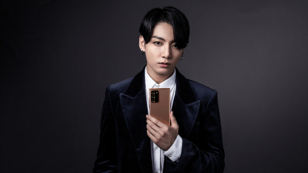 Jungkook wearing a black velvet jacket holding a smartphone against a dark background