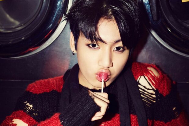 Jungkook wearing a ripped red and black sweater holding a lollipop to his lips