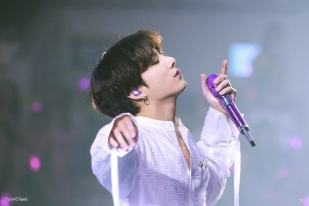 Jungkook singing on stage holding a purple microphone in a white outfit