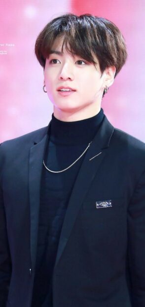 Jungkook wearing a black turtleneck and blazer with hoop earrings on a pink background