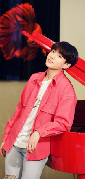 Jungkook wearing a pink jacket leaning on a red piano with a red decoration in the background