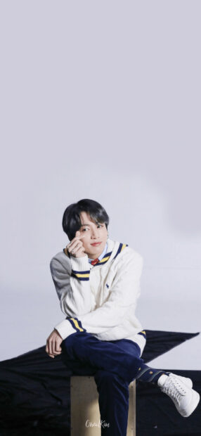 Jungkook sitting cross legged wearing a white sweater and navy pants in a relaxed pose