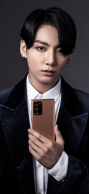 Jungkook holding a smartphone while wearing a dark velvet jacket and white shirt