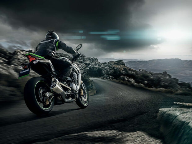 A Kawasaki motorcycle rider in full gear rides on a winding mountain road under a cloudy sky
