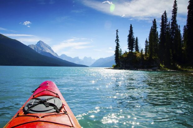 A red kayak paddling on a clear blue lake surrounded by mountains and pine trees under a bright sky