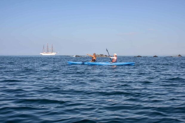 Two people kayaking on the ocean near rocks with a large sailboat in the distance