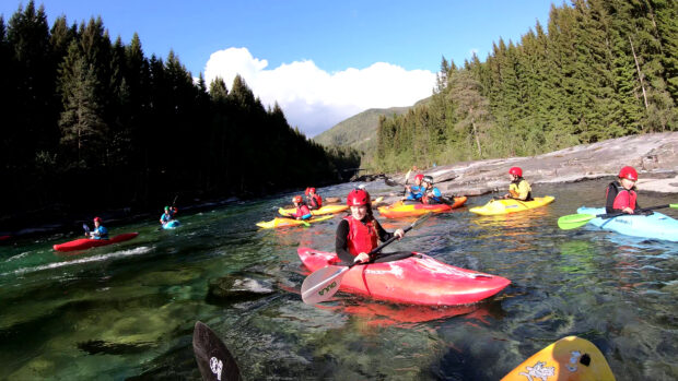 A group of kayakers paddling on a clear river surrounded by forest and mountains