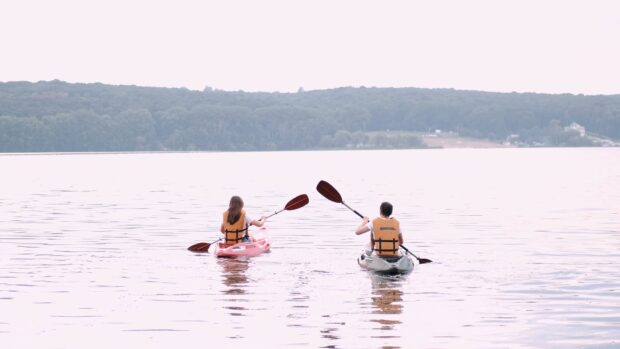 Two people kayaking on a calm lake surrounded by trees wearing orange life jackets