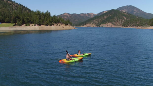 Two people kayaking on a lake surrounded by forested mountains under clear blue sky