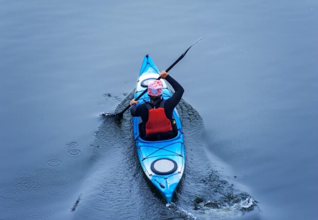 A person kayaking in a blue kayak on calm water wearing a red life vest