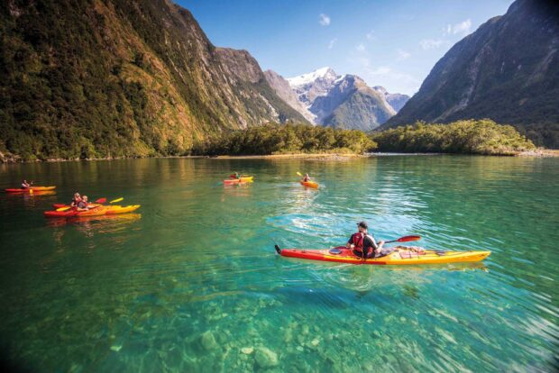 Kayakers paddling along a clear lake surrounded by mountains and forests