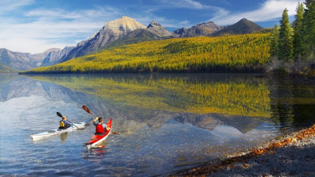 Two people kayaking on a calm lake surrounded by mountains and autumn trees