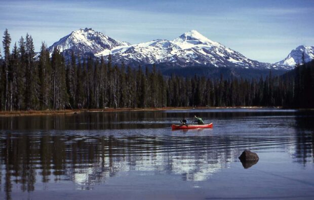Two people kayaking on a lake surrounded by forest and snow covered mountains in the background