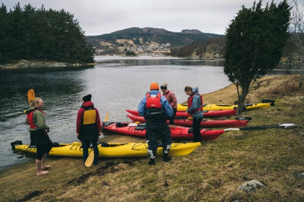 A group of people preparing kayaks on the shore near a calm lake surrounded by hills