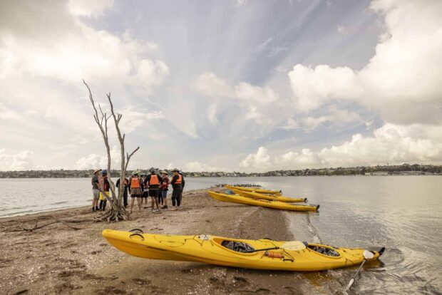 A group of people wearing life jackets preparing for kayaking on the shore with yellow kayaks nearby