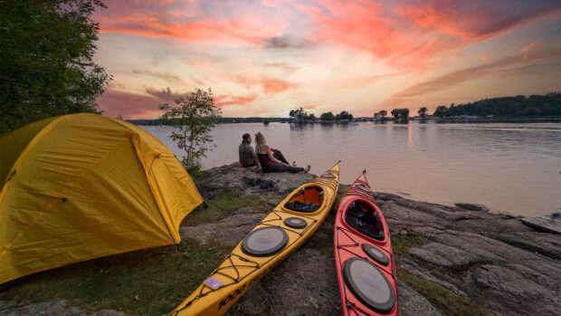A couple relaxing beside kayaks and a yellow tent near the lake at sunset