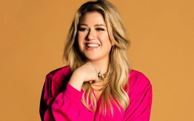 Kelly Clarkson smiling in a bright pink top with long blonde hair against a yellow background