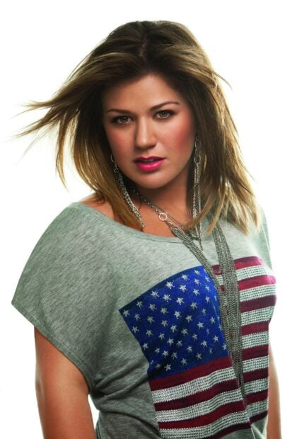 Kelly Clarkson wearing an American flag shirt and silver necklaces with bright makeup looking at the camera
