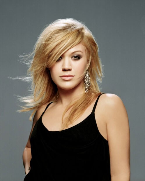 Kelly Clarkson with blonde hair wearing a black top and silver earrings against a gray background
