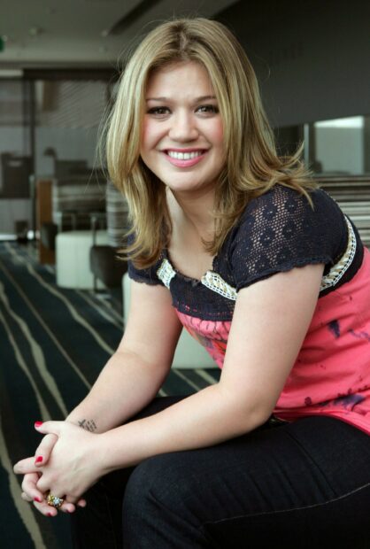 Kelly Clarkson sitting indoors wearing a pink top and smiling at the camera