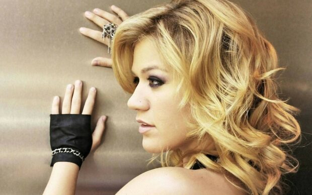 Kelly Clarkson with blonde curly hair wearing fingerless gloves against a metal background