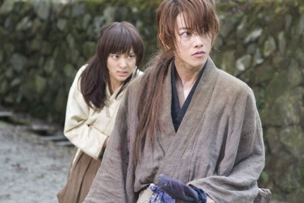 Kenshin stands alert in traditional attire with a determined expression and a woman behind him in a tense moment